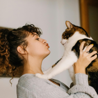 HOME 20 Mulher de cabelos cacheados em perfil fazendo bico de beijo para um gato tricolor (calico) que ela segura nos braços.