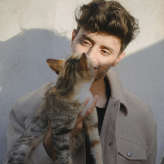 HOME 21 Homem jovem de cabelos castanhos segurando um gato malhado (tabby) próximo ao rosto em um ambiente com luz solar lateral.