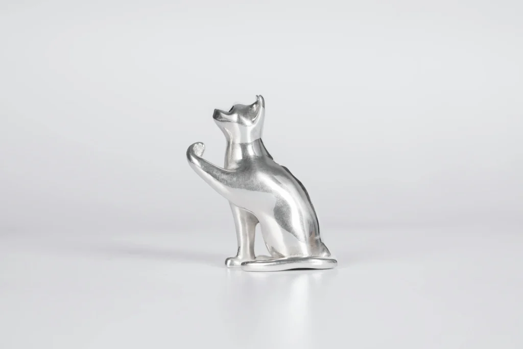 Estátua metálica elegante de um gato sentado com a pata levantada, lembrando as esculturas clássicas do Egito Antigo usadas para reverenciar os felinos como guardiões espirituais.